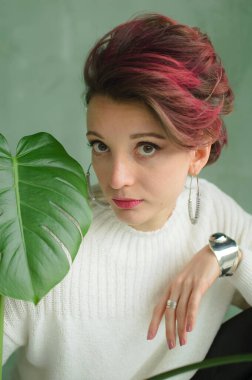 Beyaz kazaklı, gümüş küpeli, yeşil palmiye ağacının yanında oturan ve kameraya şefkatle bakan güzel bir bayan. Güzellik ve moda konsepti