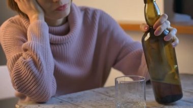 Kotlu ve pembe kazaklı üzgün kadın masada oturuyor ve bardaktan viski içiyor, koyu yeşil şişe görünür, kadın alkolizmi