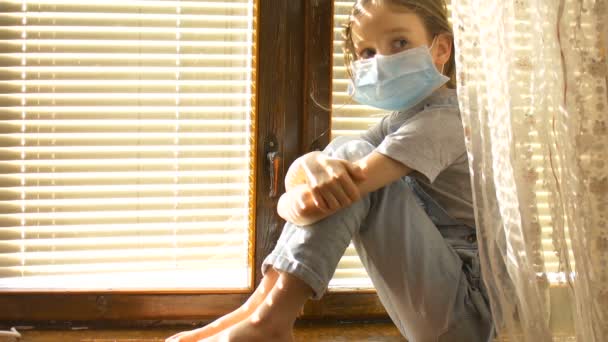 Уставшая от скуки маленькая девочка сидит на подоконнике в защитной одноразовой маске на лице во время самоизоляции дома из-за коронавируса Ковид-19 на фоне жалюзи — стоковое видео