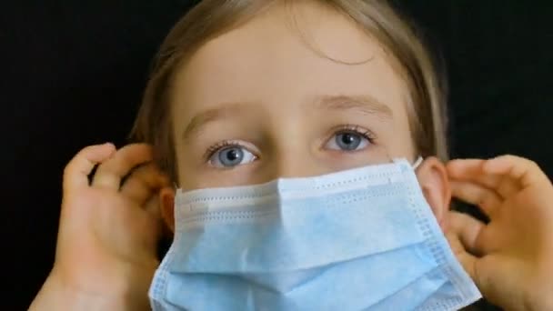 Pequena menina loira com olhos azuis e cabelos longos está posando no backgound preto em máscara cirúrgica descartável para a proteção do Coronavirus Covid-19 durante o auto-isolamento, enquanto ficar em casa — Vídeo de Stock