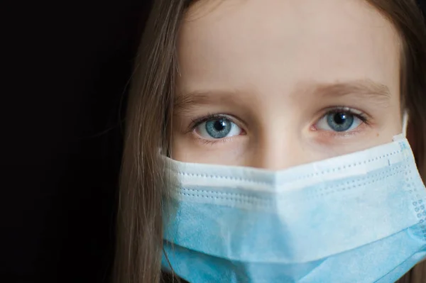 Retrato de cerca de la niña indefensa sobre fondo negro en máscara desechable azul para la protección del Coronavirus Covid-19 durante el autoaislamiento en casa — Foto de Stock