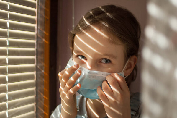 Little girl is sitting on the windowsill and trying to put protective disposable mask on her face during self-isolation at home because of Coronavirus Covid-19 on blinds background