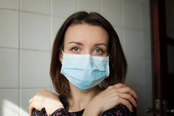 Sad adult girl staying at home because of the pandemia of Coronavirus Covid-19 and self-isolation. Woman is at the kitchen during sunny day on white tiles background in disposable mask — Stock Photo, Image