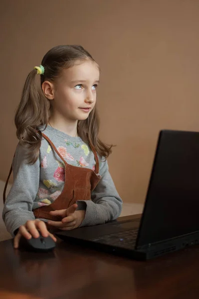 Poważna dziewczynka jest skoncentrowana przed swoim czarnym laptopem i skupiona na odrabianiu lekcji podczas kwarantanny z powodu pandemii Coronavirus Covid-19. Zdalna edukacja — Zdjęcie stockowe