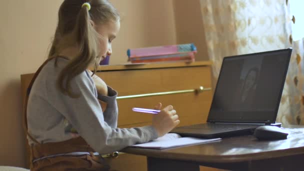 Niña rubia con dos colas de caballo estudia con su maestra que muestra tarjetas usando video chat y escribe algo en el libro de ejercicios en casa debido al auto-aislamiento debido a Covid-19 — Vídeo de stock