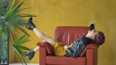 Punk rock tarzı parti kıyafetleri içinde göz kamaştırıcı bir kadın. Gayri resmi giyinmiş koyu pembe saçlı, siyah deri ceketli ve küpeli. Sarı arka planda uzun, mükemmel bacaklı kırmızı koltukta oturuyor.