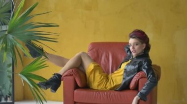 Glam rock tarzı, koyu pembe saçlı, kısa elbiseli, siyah deri ceketli, sarı arka planda kırmızı koltukta oturan ve palmiye ağacının yanında dinlenen güzel bir kıza benziyor.