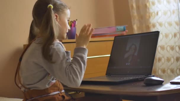 Rückansicht eines kleinen Mädchens mit zwei Pferdeschwänzen, das mit einer Videokamera auf einem schwarzen Laptop zu Hause Hallo zu ihrem Tutor sagt, weil es sich aufgrund des Coronavirus Covid-19 selbst isoliert hat. Fernausbildung — Stockvideo