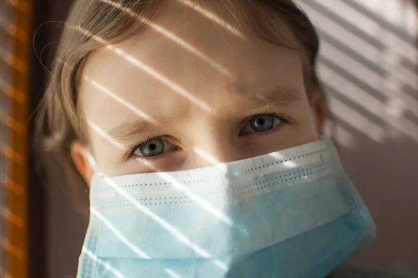 Coronavirus Covid-19 outbreak. Little blonde girl with blue eyes wearing disposable mask for protection of virus sitting near the window with blinds. Personal protective equipment — Stock Photo, Image
