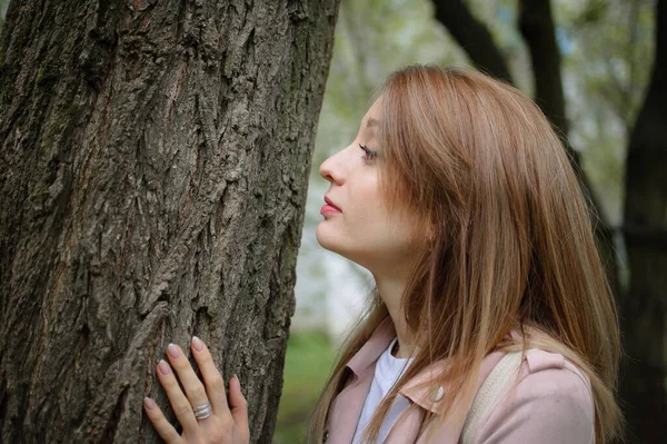 Egy fiatal, vonzó, szőke hajú nő pózol egy nagy fa mellett egy tavaszi parkban a szabadban. Egy fának támaszkodik, hogy megérintse a kérget. — Stock Fotó