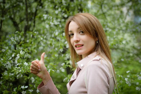 ブロンドの女性は、早春の間に庭の開花木の背景に親指を示しています。アレルギーの概念を止める — ストック写真
