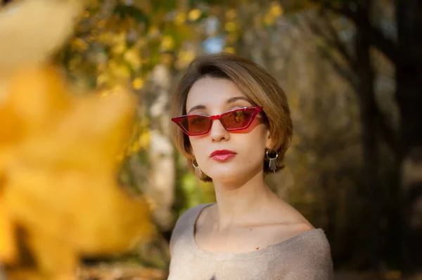 Joven y hermosa chica con pelo corto y gafas de sol rojas de moda está posando contra los árboles con hojas amarillas de fondo pasar tiempo en el parque de otoño —  Fotos de Stock