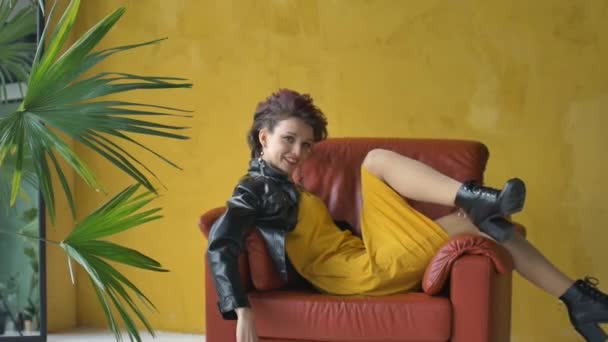Glam rock styl vzhled krásné dívky s tmavě růžové vlasy a Mohawk na sobě krátké šaty a černé kožené sako sedí v červeném křesle na žlutém pozadí v blízkosti palmy a relaxační — Stock video