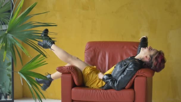 Гламурная леди в стиле панк-рок вечеринки одежда с темно-розовыми волосами в повседневной одежде, черная кожаная куртка, и серьги сидит в красном кресле с длинными идеальными ногами на желтом фоне — стоковое видео