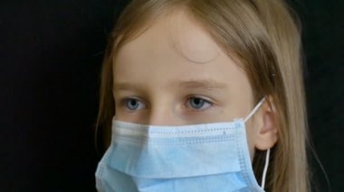 Mavi gözlü ve uzun saçlı küçük sarışın kız evde yalnızken Coronavirus Covid-19 'un korunması için ameliyat maskesiyle siyah arka planda poz veriyor.