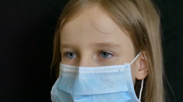 Το ξανθό κοριτσάκι με τα μπλε μάτια και τα μακριά μαλλιά ποζάρει σε μαύρο πίσω μέρος με χειρουργική μάσκα μιας χρήσης για την προστασία του Coronavirus Covid-19 κατά τη διάρκεια της αυτο-απομόνωσης, ενώ διαμένουν στο σπίτι — Αρχείο Βίντεο