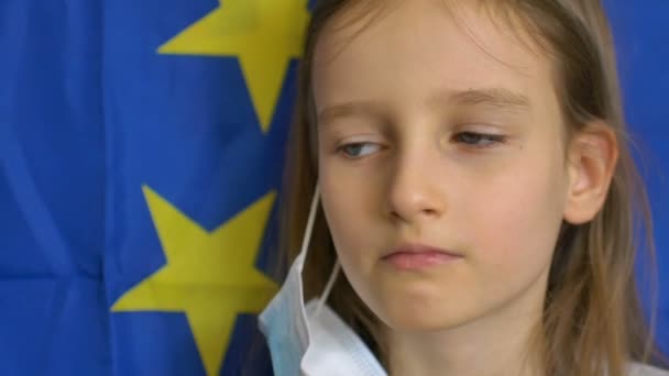 Potret close-up gadis kecil pirang dengan mata biru besar dalam topeng sekali pakai untuk perlindungan virus pada latar belakang bendera Uni Eropa di studio. Peralatan pelindung pribadi — Stok Video