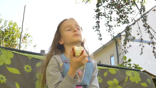 Niña rubia está comiendo manzana roja en un columpio al aire libre durante el día soleado de verano en el patio de recreo en el jardín, comida saludable, concepto de infancia feliz — Vídeo de stock