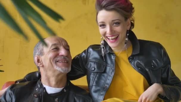 Emotional family portrait of adult daughter and senior father in loft room with houseplants. Laughing man and girl in black leather jackets in punk style on yellow wall background — Stock Video