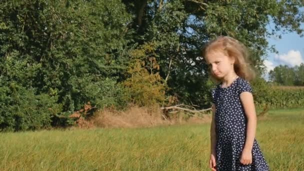 Fille bouclée blonde ennuyée est debout devant la caméra dans l'herbe verte pendant une journée. L'enfant est triste parce que les vacances d'été sont terminées et qu'il est temps de retourner à l'école — Video