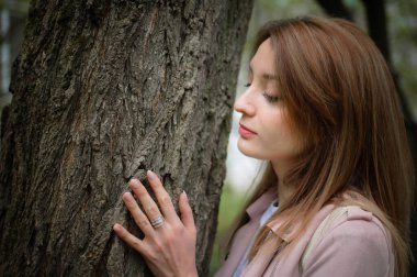 Genç ve çekici bir kadın. Sarışın. Açık hava parkında büyük bir ağacın yanında poz veriyor. Ağaca yaslanır ve kabuğuna dokunur.