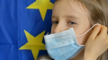Avrupa Birliği bayrak arka planında virüsü korumak için maske takan büyük mavi gözlü küçük sarışın kızın yakın plan portresi stüdyoda. Kişisel koruma ekipmanları