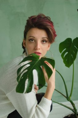 Beyaz kazaklı, gümüş küpeli, yeşil palmiye ağacının yanında oturan ve kameraya şefkatle bakan güzel bir bayan. Güzellik ve moda konsepti