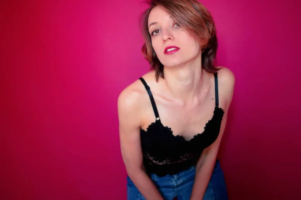 Young woman with short hair dressed in black lace bustier and blue jeans is posing in the studio on bright pink background