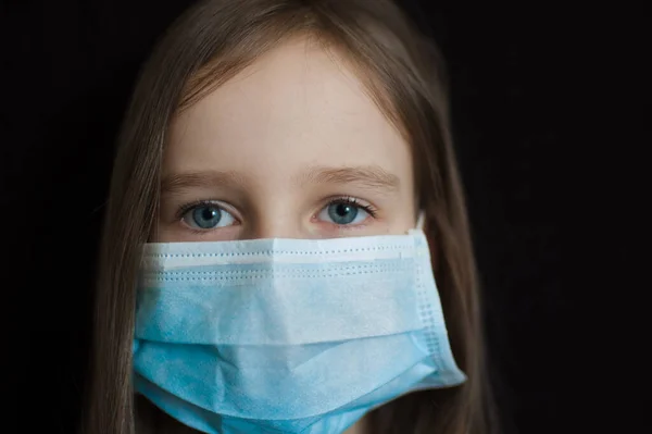 Retrato de chica triste con los ojos azules está mirando a la cámara en el fondo negro en la máscara quirúrgica desechable para la protección del Coronavirus Covid-19 durante el auto-aislamiento durante su estancia en casa —  Fotos de Stock