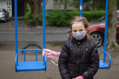 Kapalı oyun parkında gri maskeli ve koyu renk ceketli üzgün kızın portresi. COVID-19 'a karşı karantina önlemleri. Koronavirüs salgını.