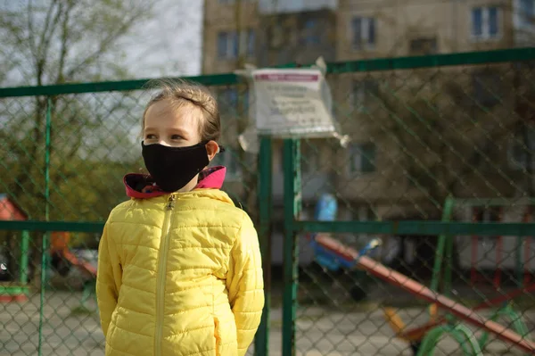 Sad little girl in face mask in yellow jacket standing near closed playground outdoor. Kids play area locked because of Coronavirus social distance quarantine.