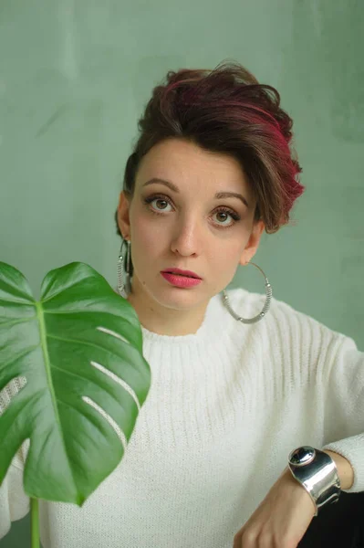 Schöne Dame in weißem Pullover, silbernen Ohrringen und Armreif sitzt neben einer grünen Palme und blickt zärtlich in die Kamera. Schönheits- und Modekonzept — Stockfoto