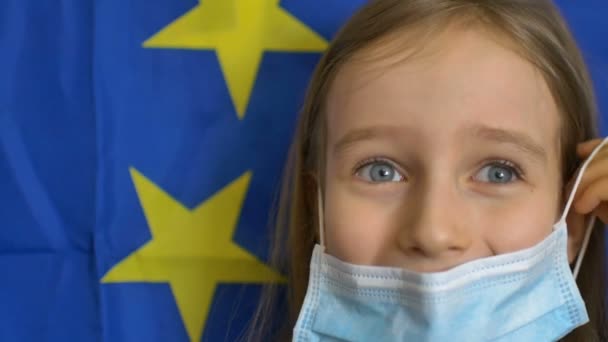 Zbliżenie portret blondynki z dużymi niebieskimi oczami w masce do ochrony przed wirusem na banerze flagowym Unii Europejskiej w studio. Środki ochrony indywidualnej — Wideo stockowe
