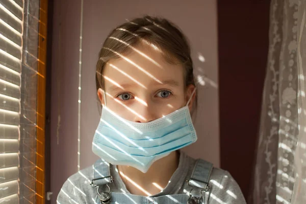 Brote de Coronavirus Covid-19. Niña rubia con ojos azules usando máscara desechable para la protección del virus sentado cerca de la ventana con persianas. Concepto de equipo de protección individual — Foto de Stock