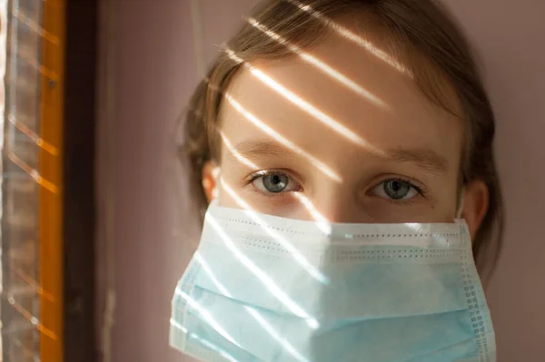 Brote de Coronavirus Covid-19. Niña rubia con ojos azules usando máscara desechable para la protección del virus sentado cerca de la ventana con persianas. Equipos de protección individual — Foto de Stock