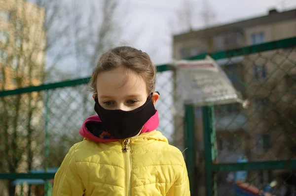 Niña triste en máscara facial en chaqueta amarilla de pie cerca de patio cerrado al aire libre. Área de juegos para niños bloqueada debido a la cuarentena de distancia social por Coronavirus . —  Fotos de Stock