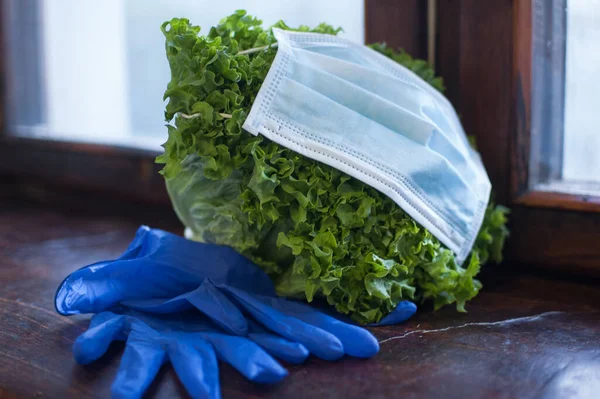 Green leaves of the salad in surgical mask lying on the wooden windowsill near blue protective gloves. Proper nutrition, delivery food during a Coronavirus pandemic and isolation.