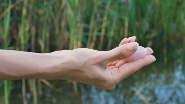 Female hand holding a rose quartz crystal yoni egg on river background. Womens health, unity with nature concepts — Stock Video