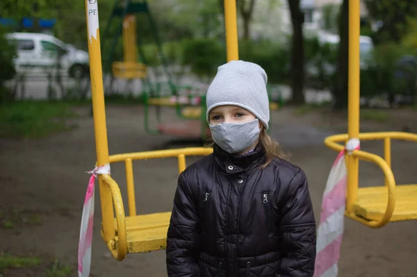 在室外密闭的操场上,一个身穿灰色面具和深色夹克的忧伤女孩的画像.对COVID-19采取检疫措施。Coronavirus大流行病. — 图库照片