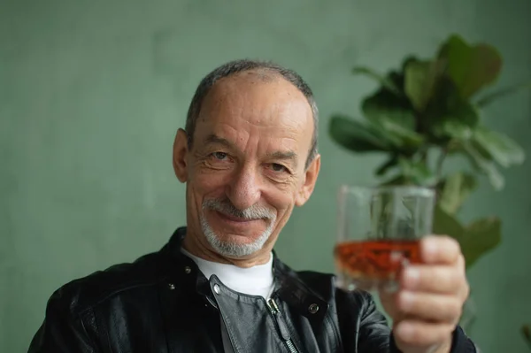 Вид спереди пожилого человека, держащего стакан виски или виски и смотрящего на камеру в комнате в стиле лофт со светло-зелеными стенами и домашними растениями на заднем плане. Алкогольная зависимость — стоковое фото
