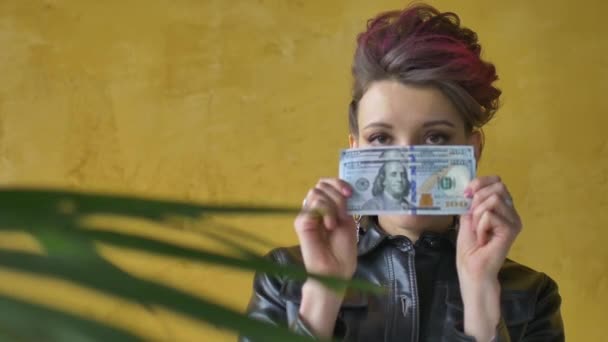 Glamorous aggressive lady in punk rock style party clothes with dark pink hair in black leather jacket and long earrings on yellow background with money dollars and hryvnias in hands — Stock Video