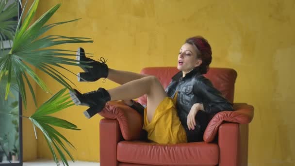 Глэм рок стиль взгляд красивой девушки с темно-розовыми волосами и ирокезом в коротком платье и черная кожаная куртка сидит в красном кресле на желтом фоне рядом с пальмой и расслабляющий — стоковое видео