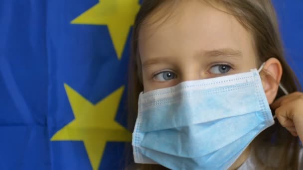 Retrato de cerca de la niña rubia con grandes ojos azules en máscara imposible para la protección del virus en el fondo de la bandera de la Unión Europea en el estudio. Equipos de protección individual — Vídeos de Stock