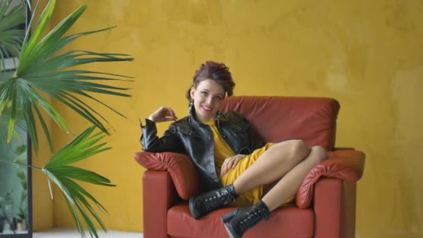Glam Rock Look eines schönen Mädchens mit dunkelrosa Haaren und Mohawk trägt kurzes Kleid und schwarze Lederjacke sitzt im roten Sessel auf gelbem Hintergrund in der Nähe der Palme und entspannt — Stockvideo