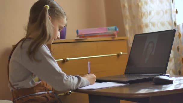Liten blond flicka med två hästsvansar studier med sin lärare med videochatt och skriva ner något i träningsbok hemma på grund av den isolering på grund av Coronavirus Covid-19 — Stockvideo