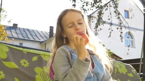 Petite fille blonde enfant mange pomme rouge sur une balançoire en plein air pendant la journée ensoleillée d'été sur l'aire de jeux dans le jardin, nourriture saine, concept d'enfance heureuse — Video