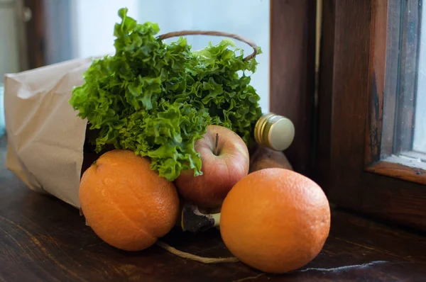 Produktset, grüner Salat und Früchte liegen in Bastelpapierverpackung auf der hölzernen Fensterbank. Lieferung von Lebensmitteln, richtiges Ernährungskonzept — Stockfoto