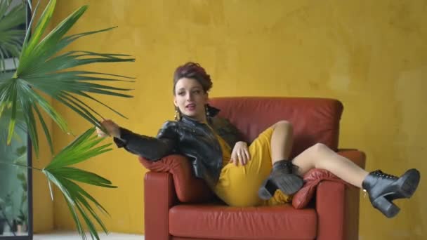 Glam rock tarzı, koyu pembe saçlı, kısa elbiseli, siyah deri ceketli, sarı arka planda kırmızı koltukta oturan ve palmiye ağacının yanında dinlenen güzel bir kıza benziyor. — Stok video