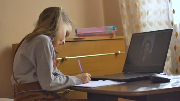 Egy szőke kislány két lófarkas tanulmánnyal a tanárával videochat-et használva, és leírni valamit otthon a füzetbe a Coronavirus Covid-19 miatti elszigeteltség miatt. — Stock videók
