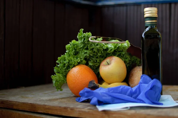 Product set on the wooden windowsill. Green leaves of the salad and fruits lying near surgical mask and blue protective gloves. Delivery food during a Coronavirus pandemic and isolation.